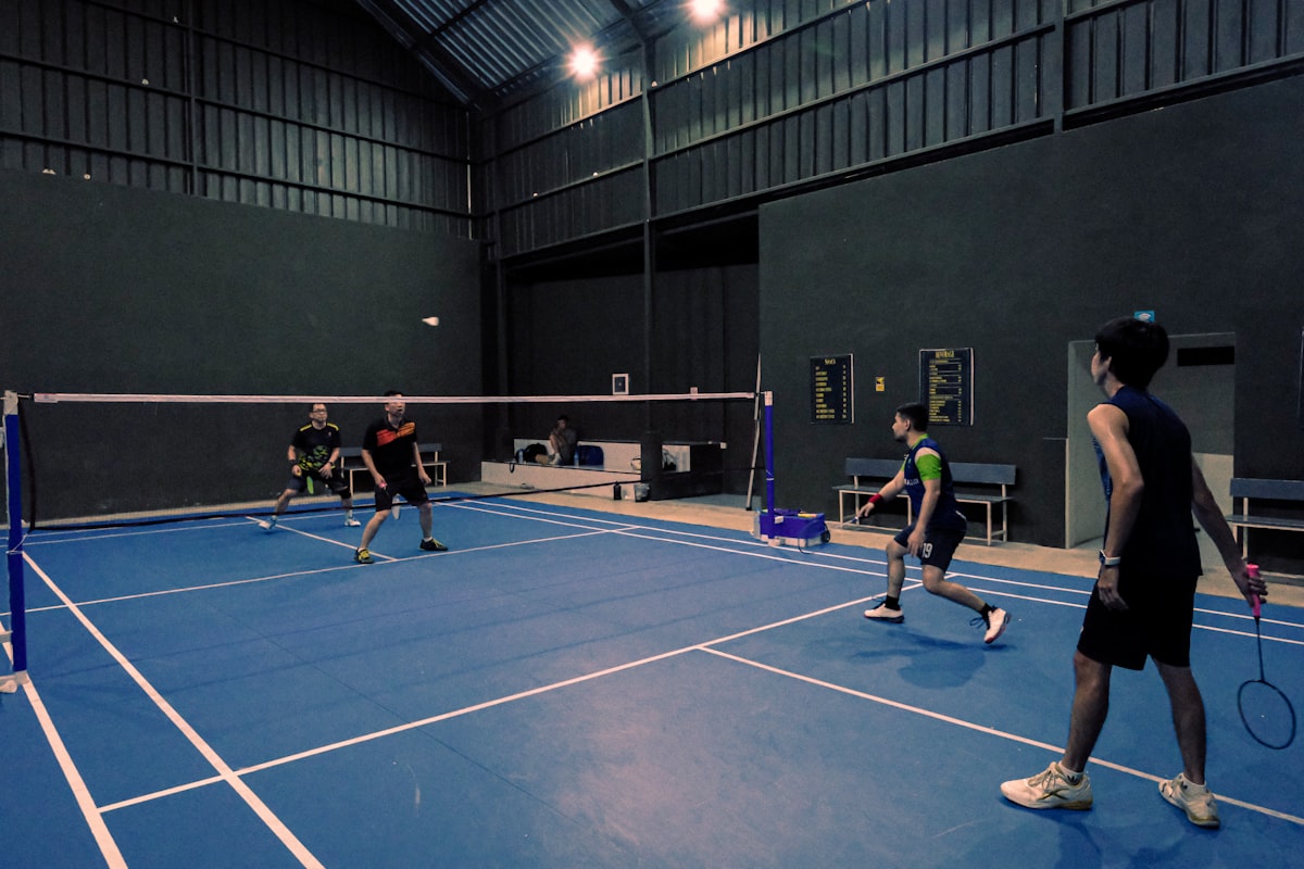 Four players in a doubles badminton match on an indoor court, one pair at the net and the other pair back, with the shuttle in mid-flight