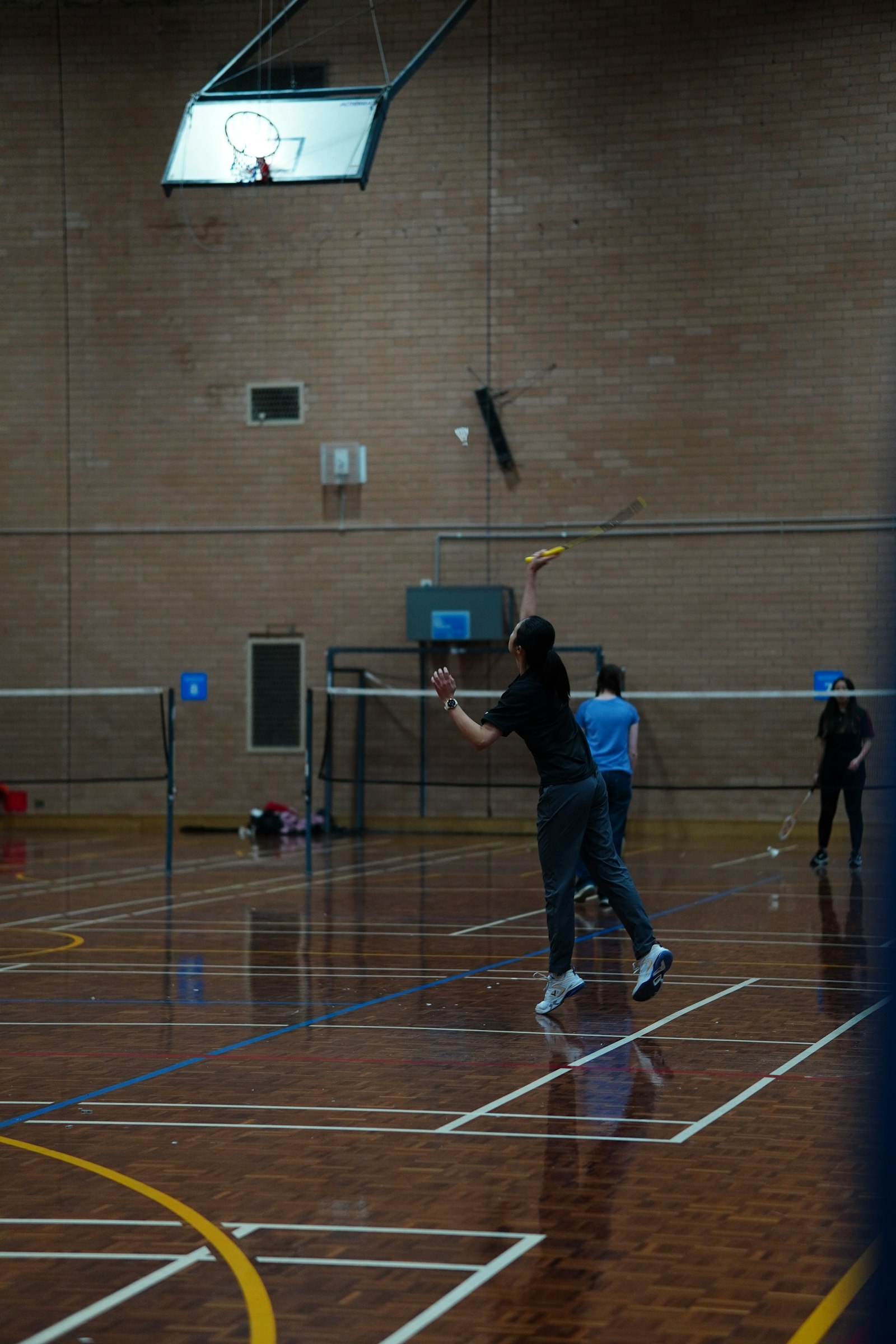 Recreational badminton players on an indoor wood-floor court, one player mid-serve, others rallying on adjacent courts in a shared gym hall