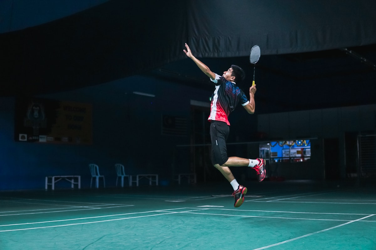 A badminton player mid-jump-smash on an indoor court, racket raised overhead with a clear view of the forehand grip on the handle