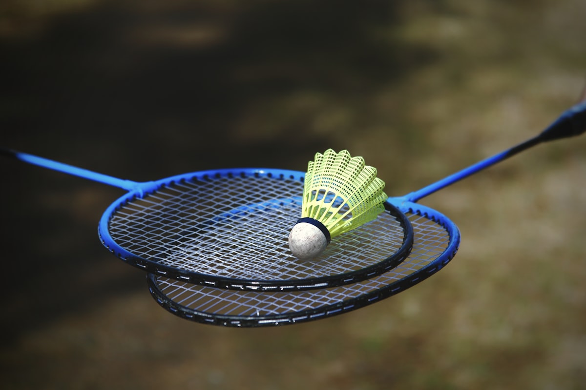 Dos raquetas de bádminton cruzadas al aire libre con un volante amarillo apoyado sobre las cuerdas