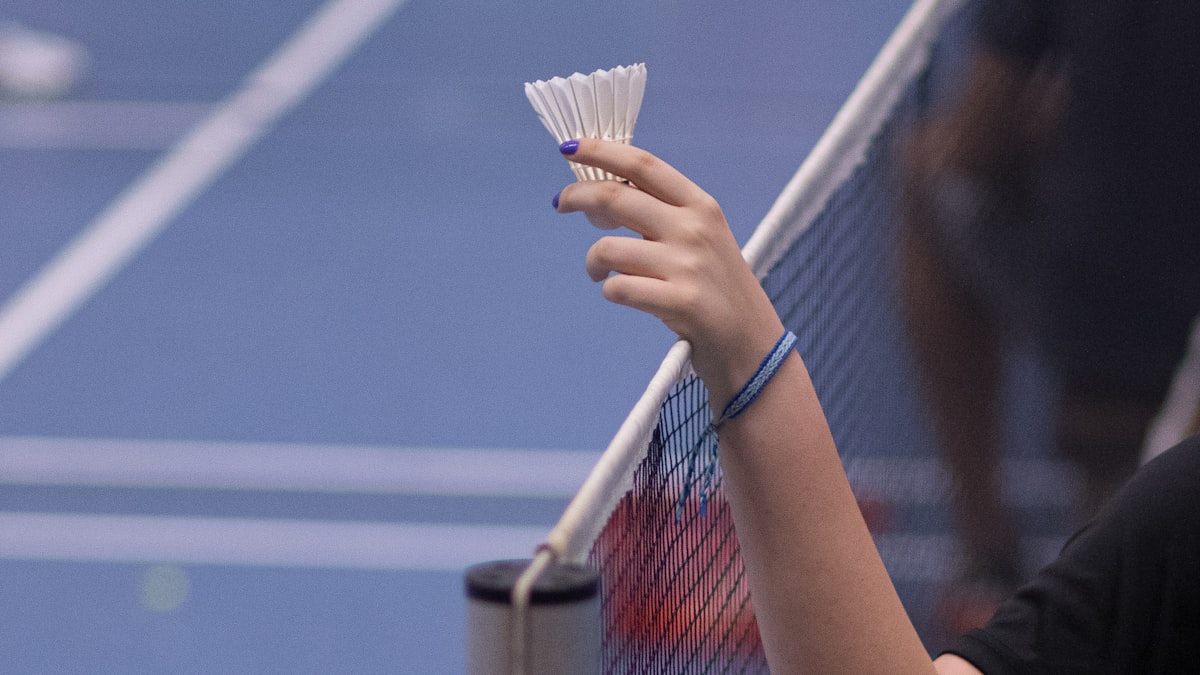 La mano de un jugador sosteniendo un volante sobre la red en una pista de bádminton cubierta, con las líneas del campo visibles