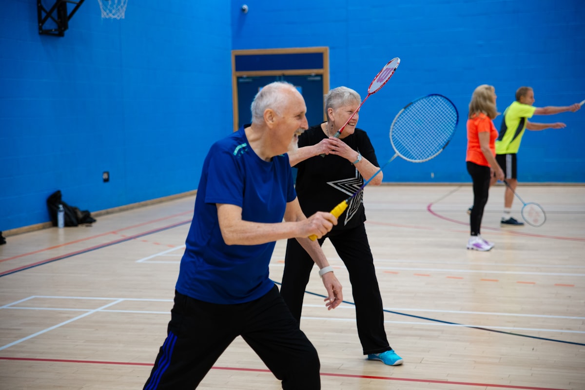 Two recreational players in a ready stance with rackets raised on an indoor gymnasium badminton court, with two more players rallying in the background
