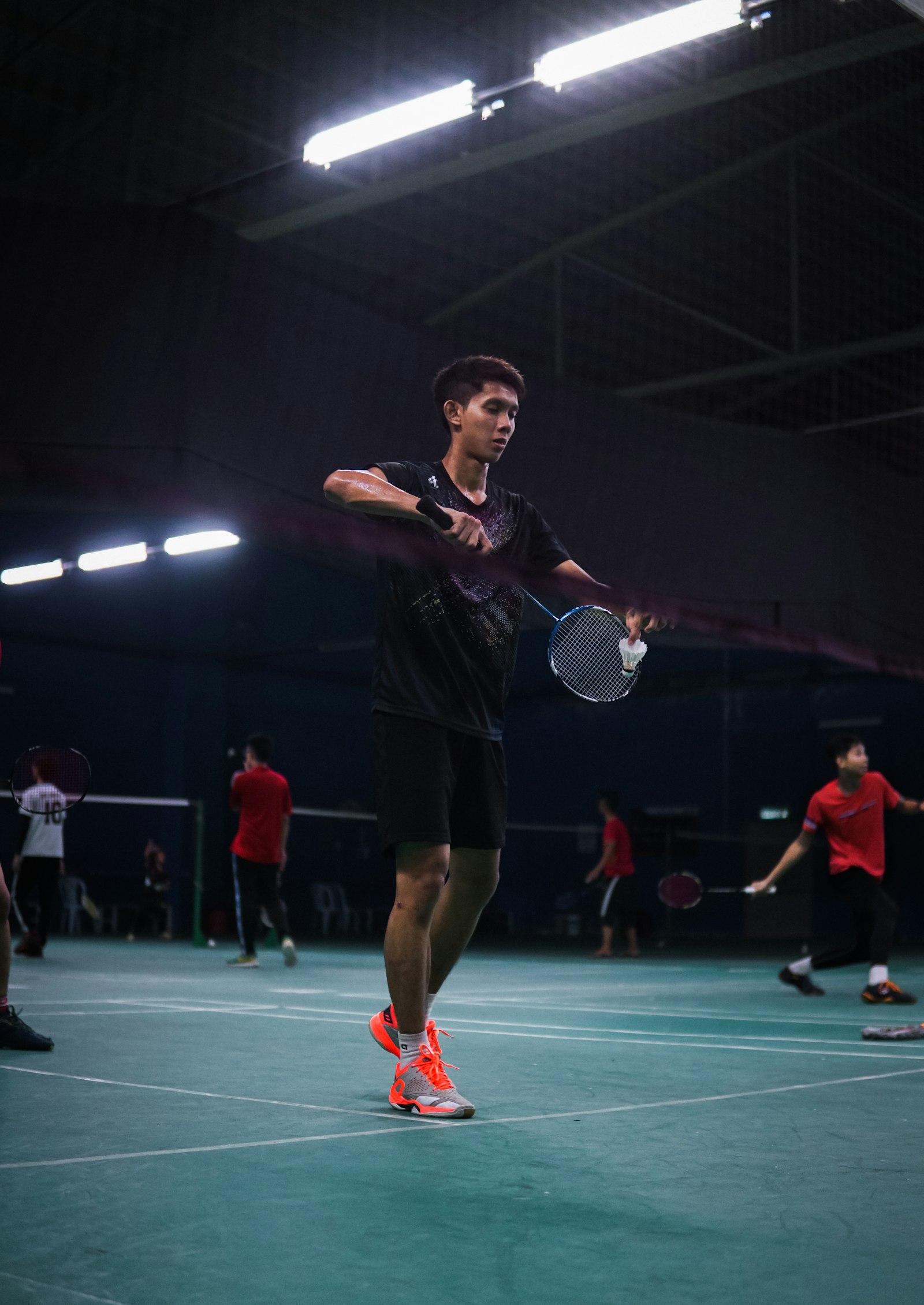 A single badminton player at the net in a recreational club match, racket extended toward the shuttle, with other players rallying on adjacent courts in the background