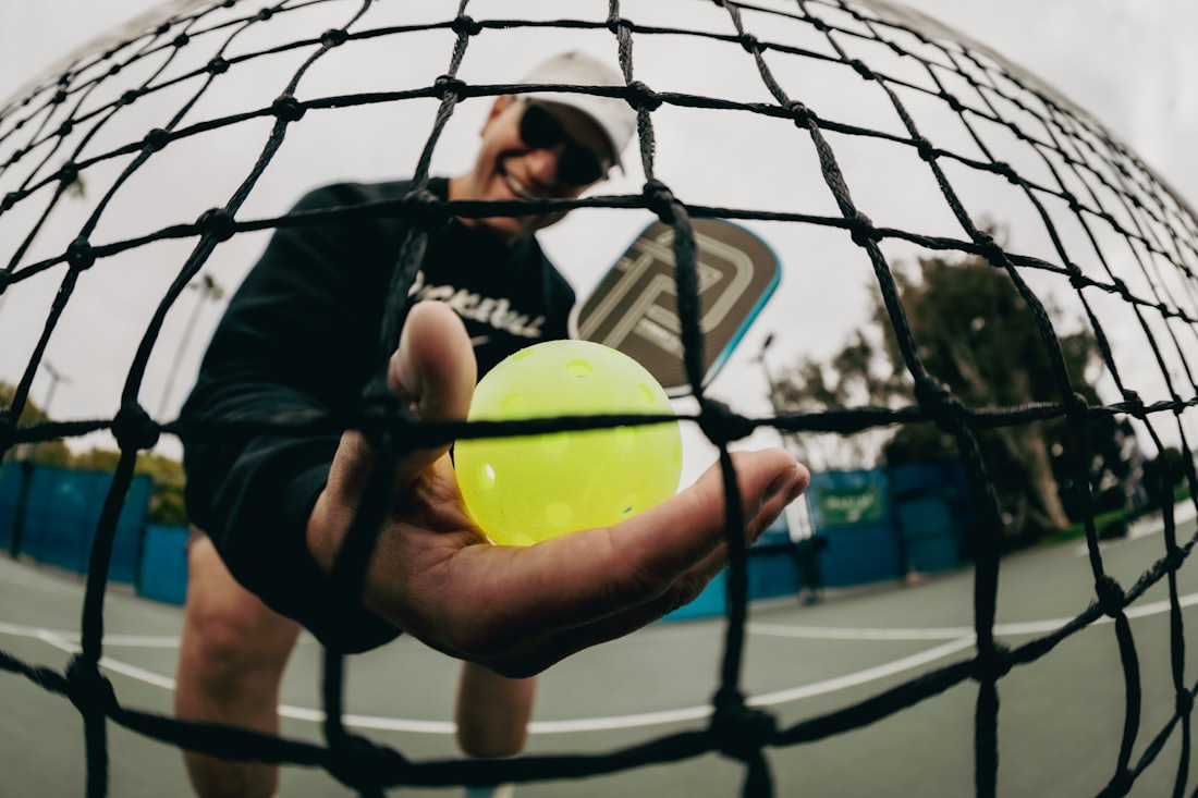 Una jugadora de pickleball sostiene una pelota amarilla de exterior con la red y la pista visibles a su espalda