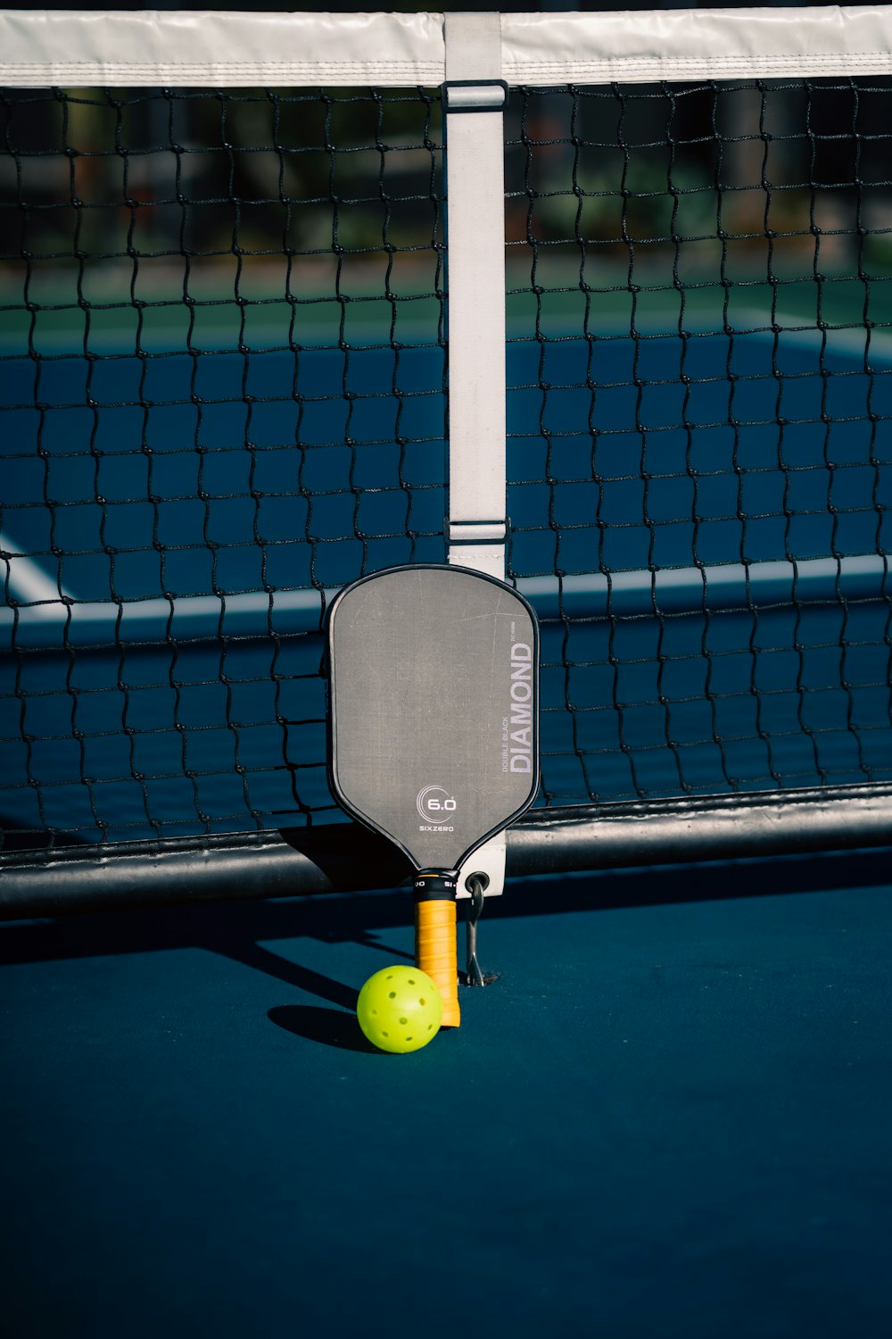 Vista a ras de pista de una pelota de pickleball junto a una pala sobre una pista recién pintada en azul y verde