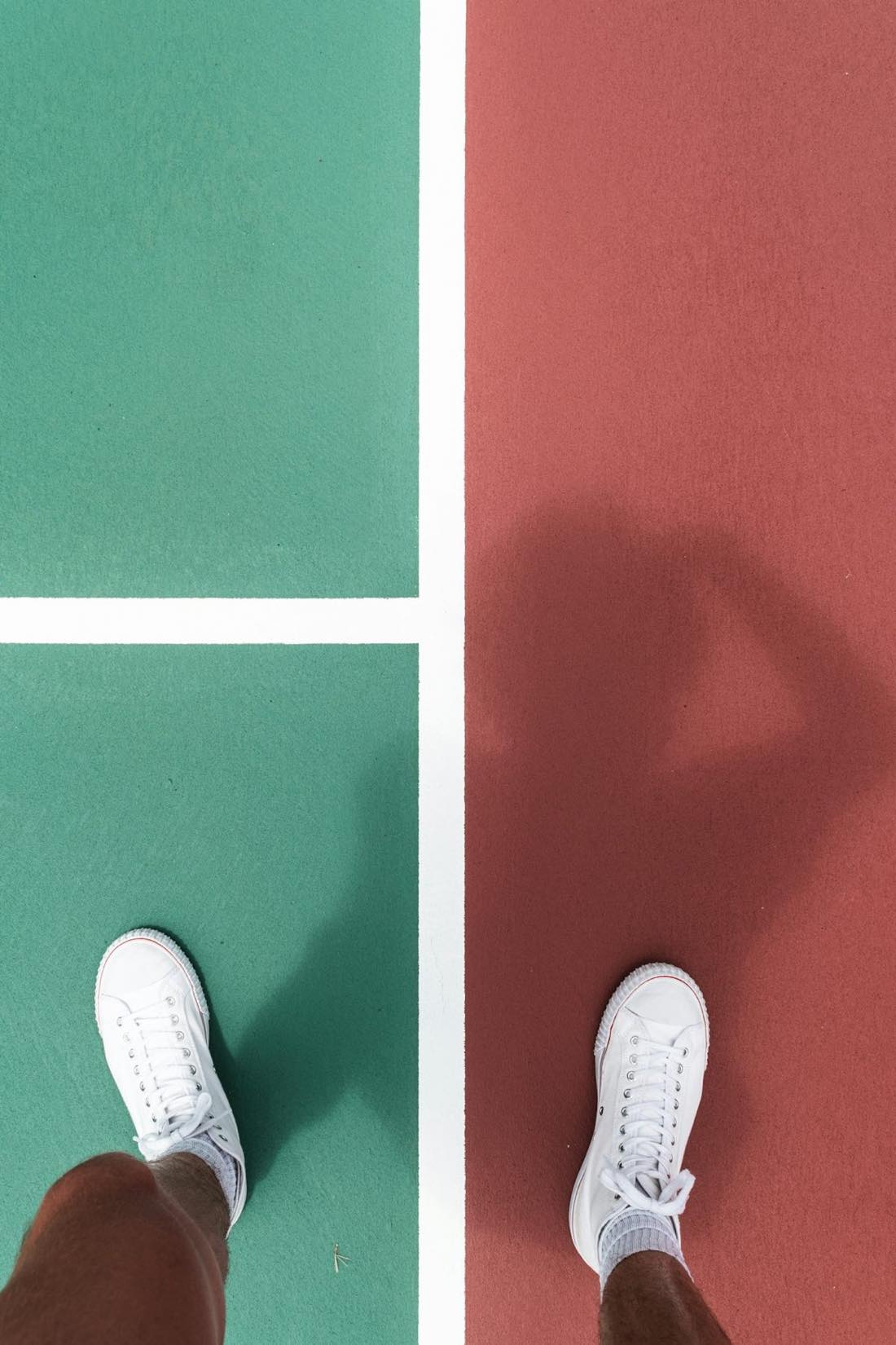 White court shoes standing on the boundary between the green and red painted surfaces of an outdoor tennis court
