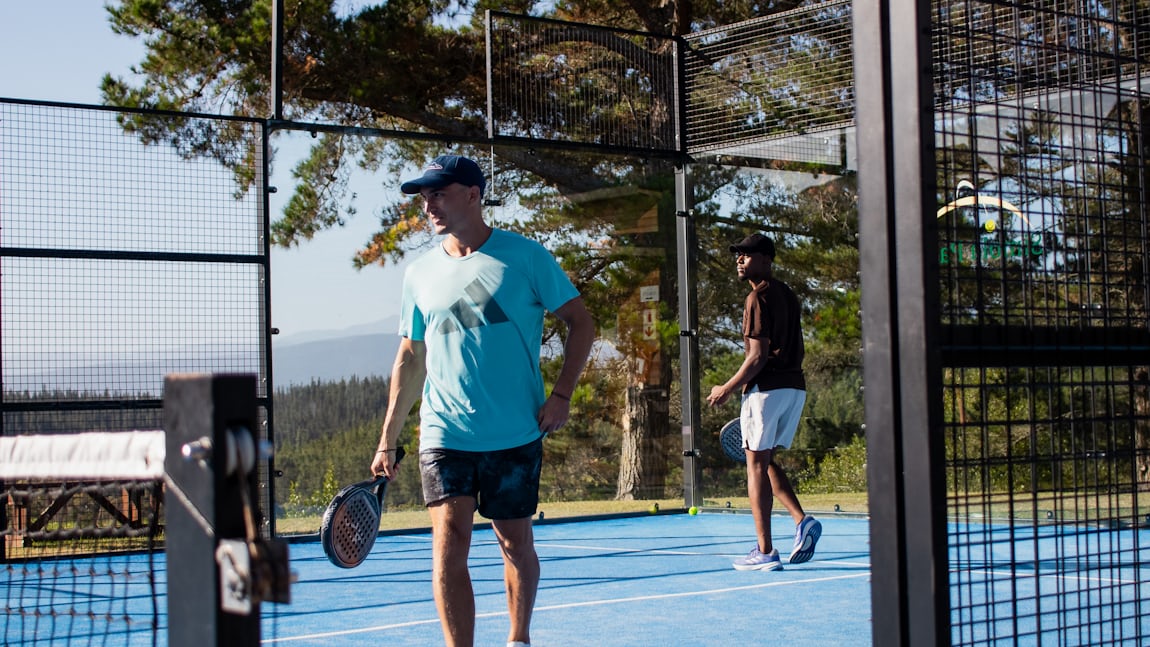 Two players on an outdoor padel court, the mesh-fence panels, glass wall section, and net-post hardware of the enclosure clearly visible.
