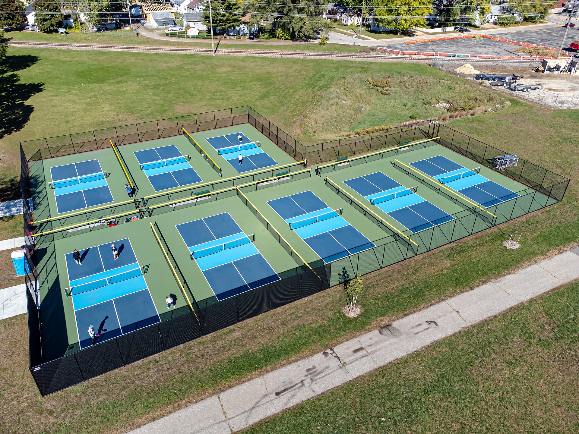 Aerial view of eight blue and green public pickleball courts inside one fenced park enclosure in La Crosse, Wisconsin, with a few players on the courts