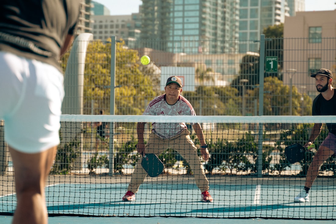 A pickleball player at the net focused on a return shot during a doubles rally, with a downtown skyline behind the court
