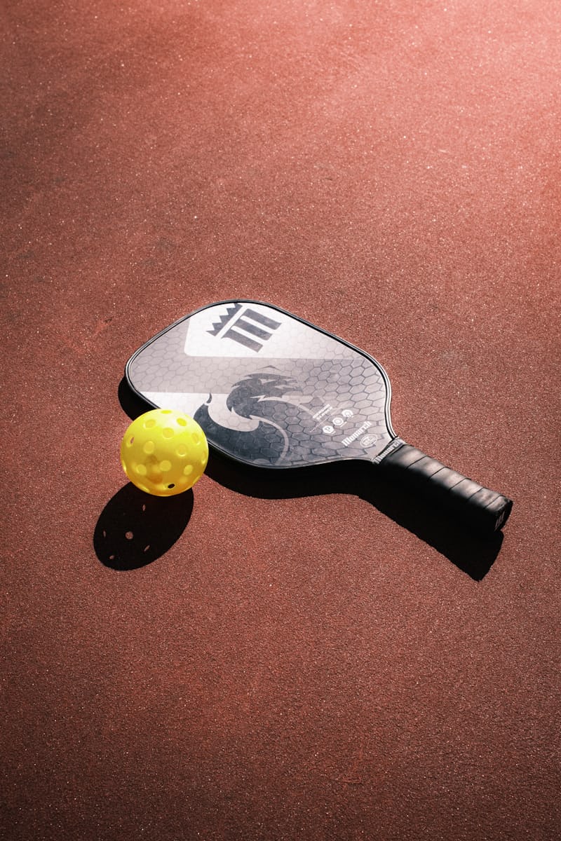 A pickleball paddle and a single yellow perforated pickleball resting on an outdoor court in warm sunlight