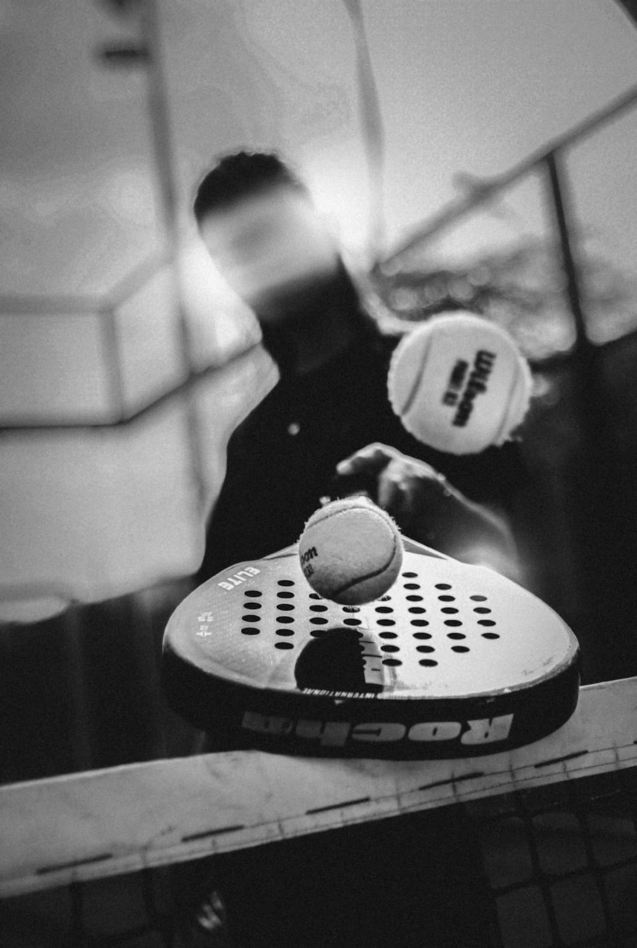 Primer plano en blanco y negro de la cara maciza y perforada de una pala de pádel golpeando una pelota en la red, con un jugador desenfocado en movimiento detrás