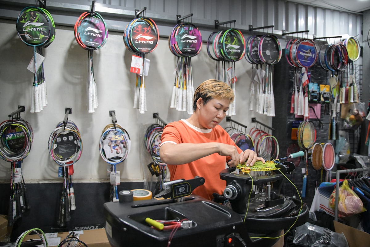 Un empleado de una tienda de deportes usando una máquina encordadora para reencordar una raqueta de bádminton, rodeado de raquetas expuestas