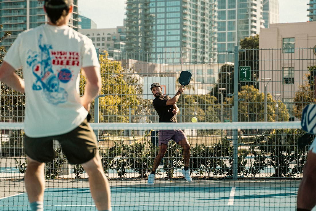 Two pairs of pickleball players rallying at a public community court complex in San Diego, with downtown high-rises in the background