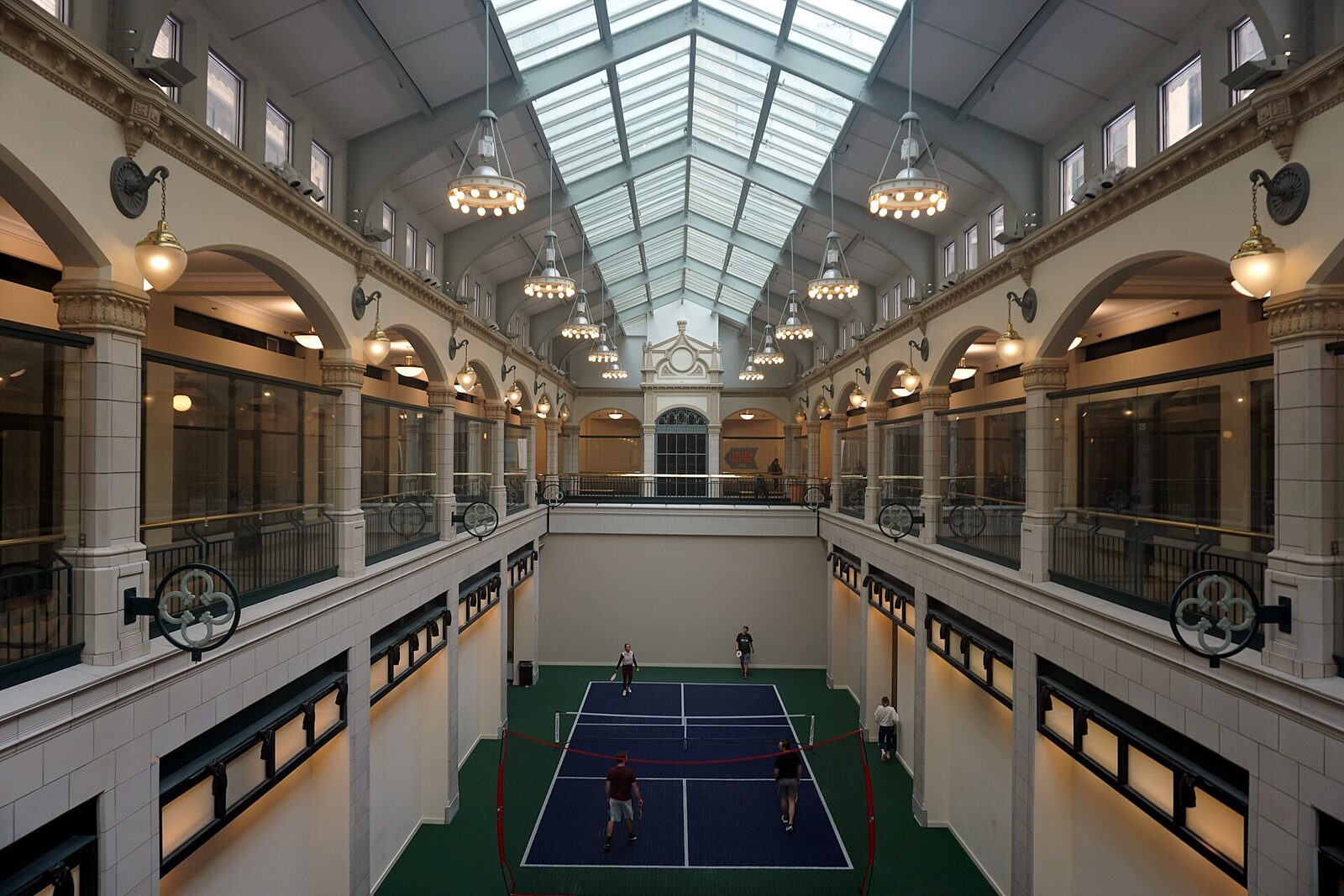 Four players on a blue pickleball court on the green floor of a glass-roofed historic arcade atrium in Milwaukee, seen from an upper balcony