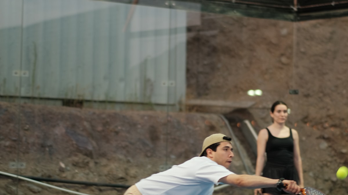 A padel player stretches for a low ball at the glass wall while a partner holds position a few steps behind — the court coverage doubles pairs manage together.