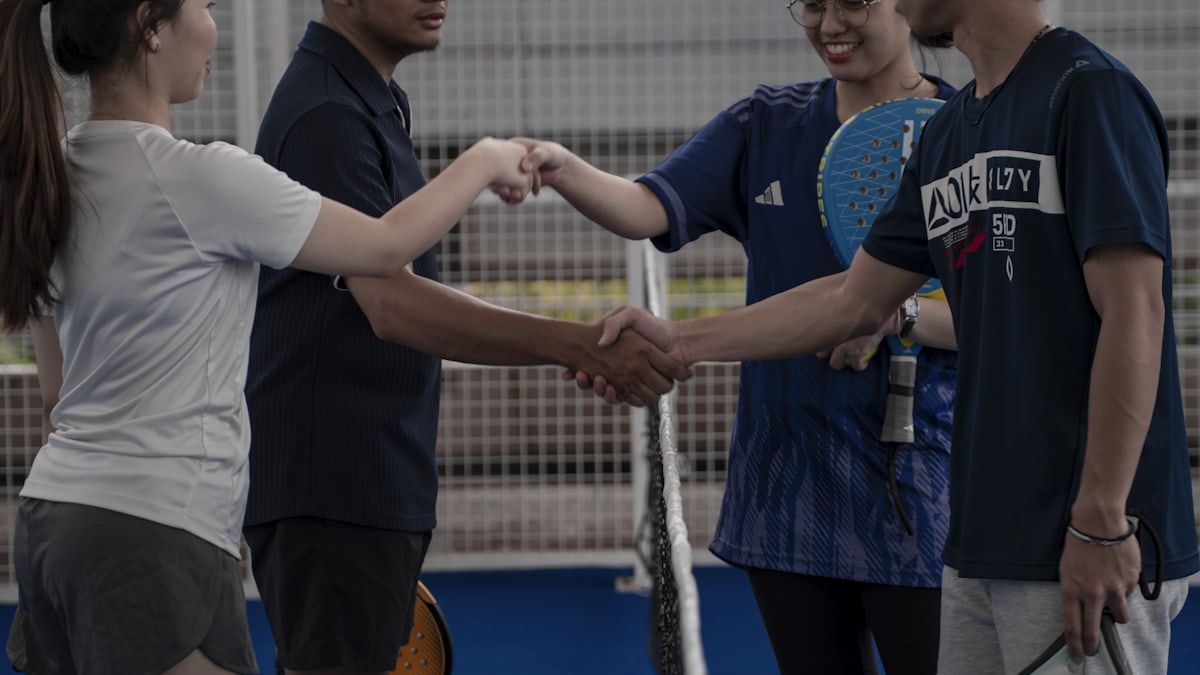 Four padel players, two per side, shake hands and fist-bump across the net after a club match, the mesh-fenced court behind them.