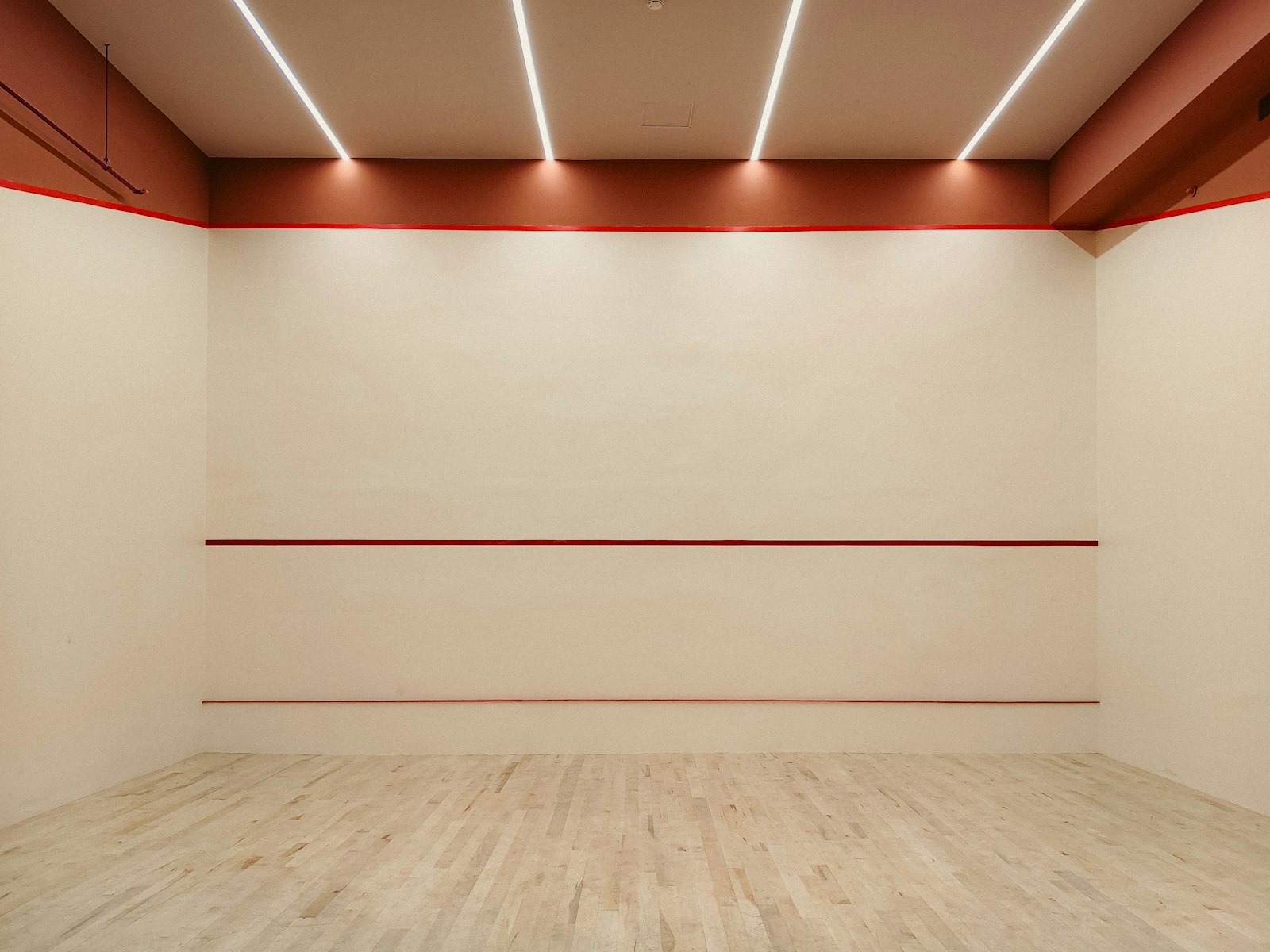 An empty squash court, front wall and out-of-court line visible, wood floor and red tin markings — the enclosed, walled-court design squash and padel share in spirit but build very differently