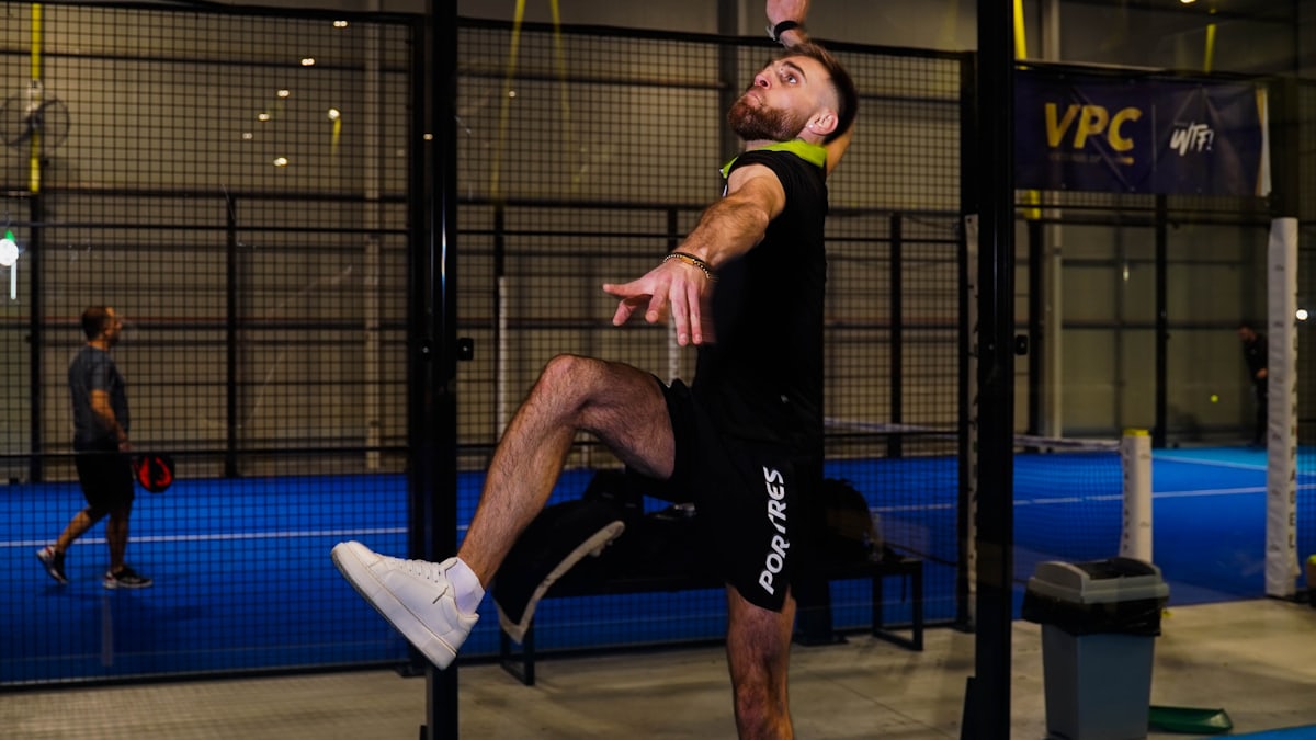A padel player leaps into an overhead smash on an indoor court, the mesh fence and net post clearly visible behind him.