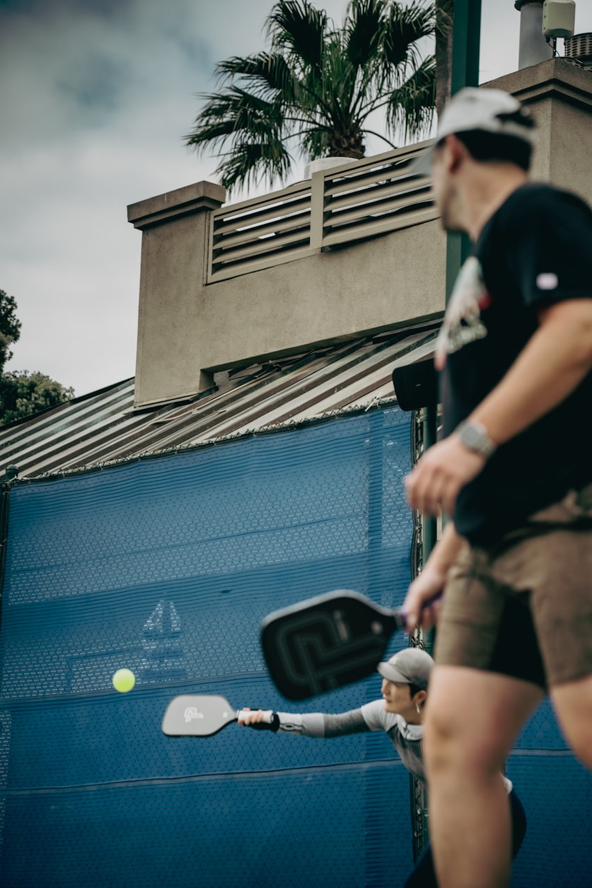 Players in mid-rally on a sunlit outdoor pickleball court with paddles and balls in motion