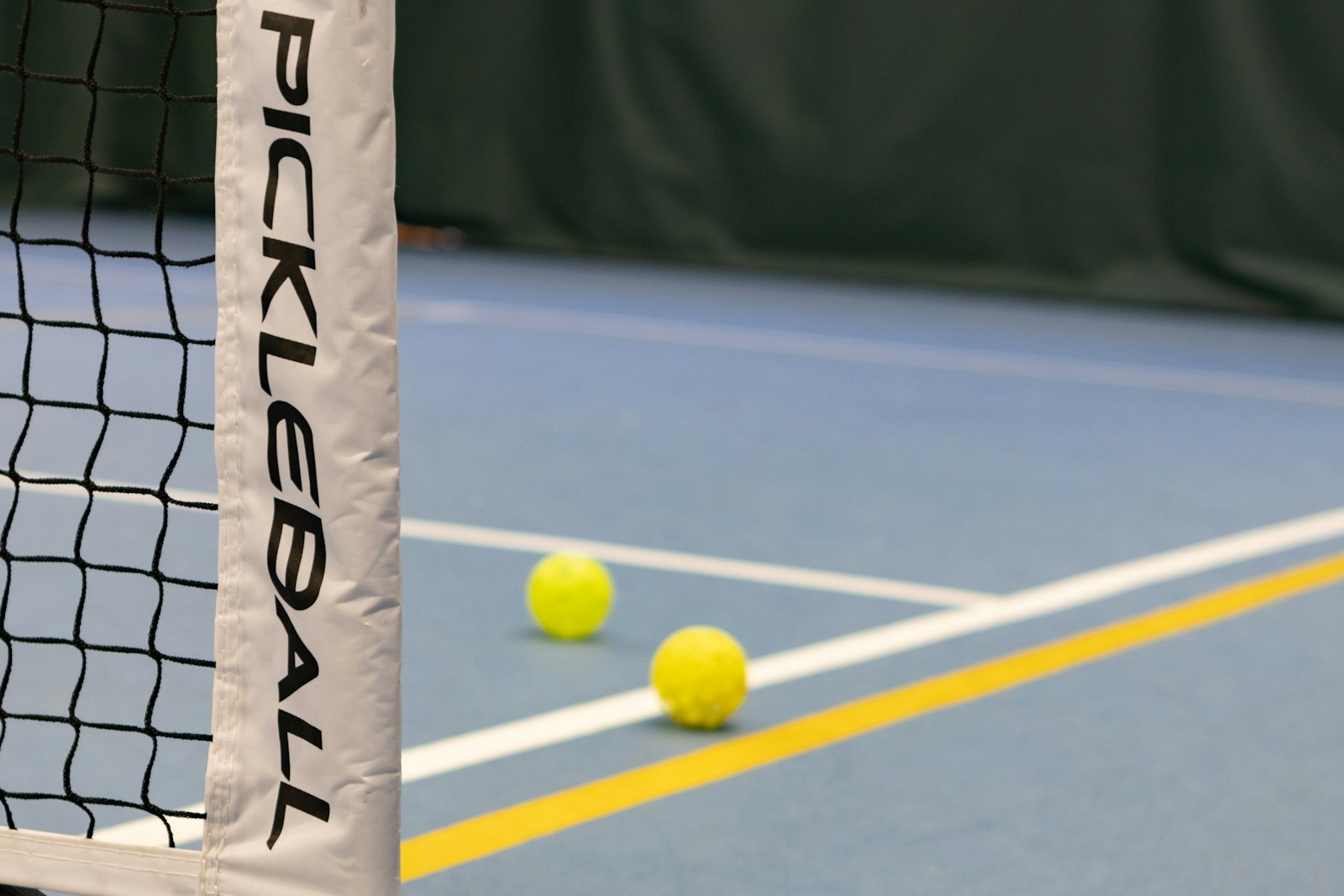 Two yellow perforated pickleballs resting on an indoor court beside a net post labeled PICKLEBALL