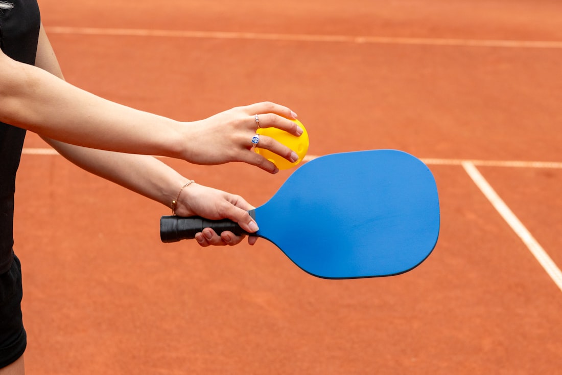 A player grips a pickleball paddle and ball at the baseline of a sunlit outdoor court