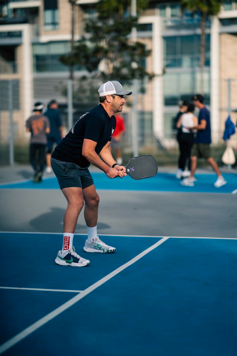 A pickleball player crouches in ready position right at the court's sideline, paddle up, other players visible behind