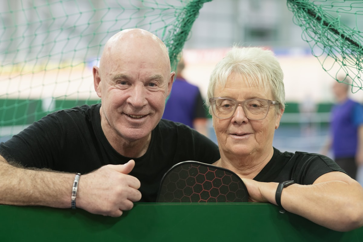 An older man and woman smiling together at the net, the woman holding a pickleball paddle, on an indoor pickleball court
