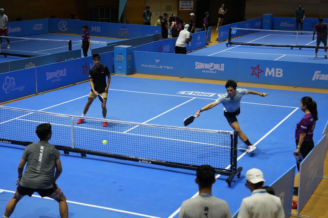 Un jugador de pickleball se lanza en una zancada completa junto a la red para alcanzar una pelota baja — el tipo de movimiento lateral rápido y extendido asociado a caídas y lesiones de tendón de Aquiles