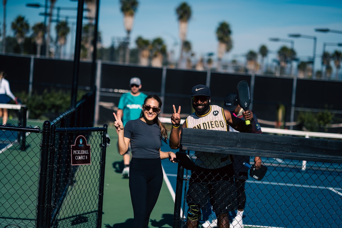 A group of smiling pickleball players flashing peace signs at the net of a multi-court facility lined with palm trees
