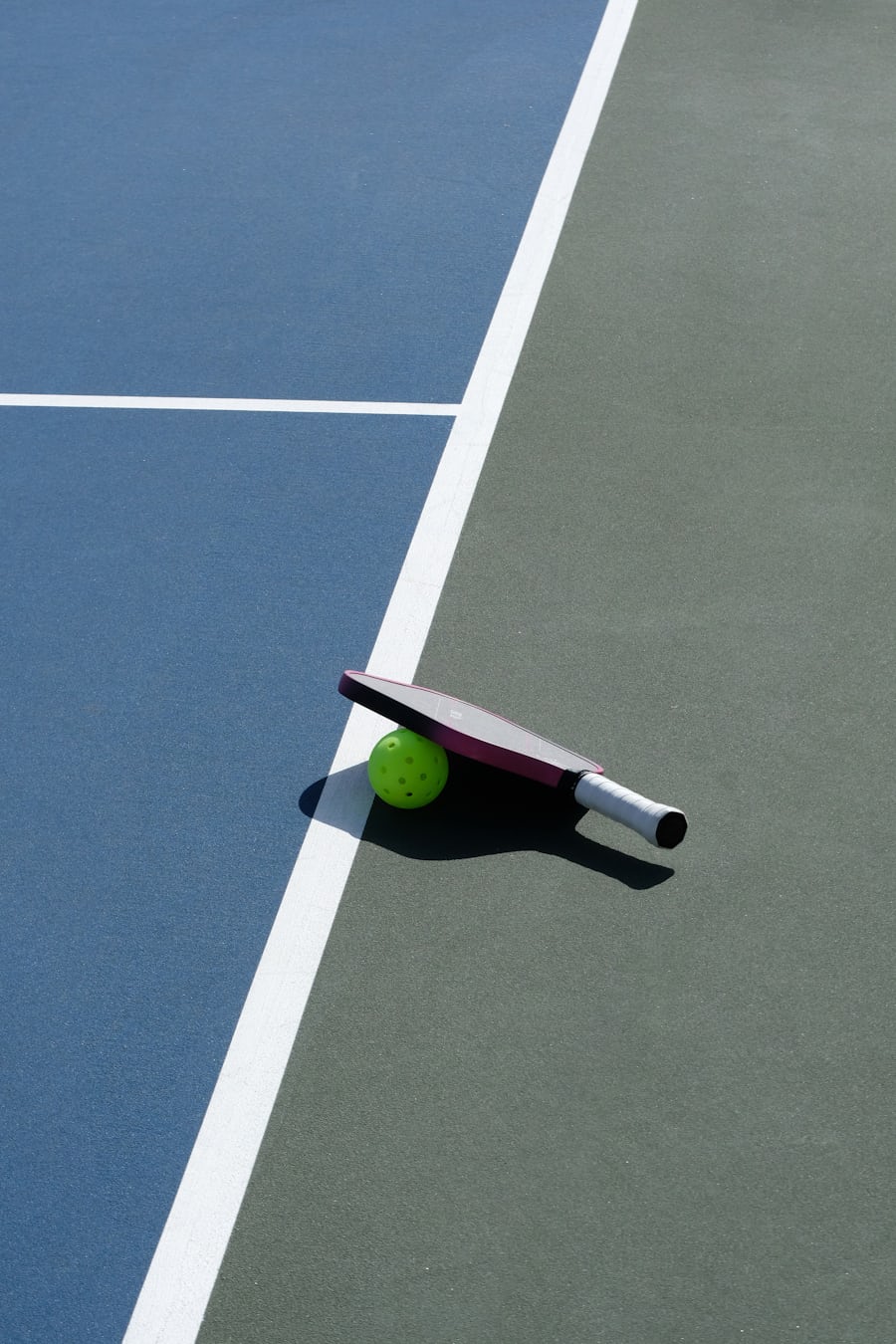 Una pala y una pelota de pickleball sobre una pista exterior soleada, proyectando una sombra marcada bajo el sol del mediodía