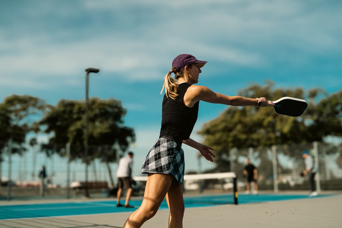A pickleball player follows through on a forehand shot with other players and courts visible in the background