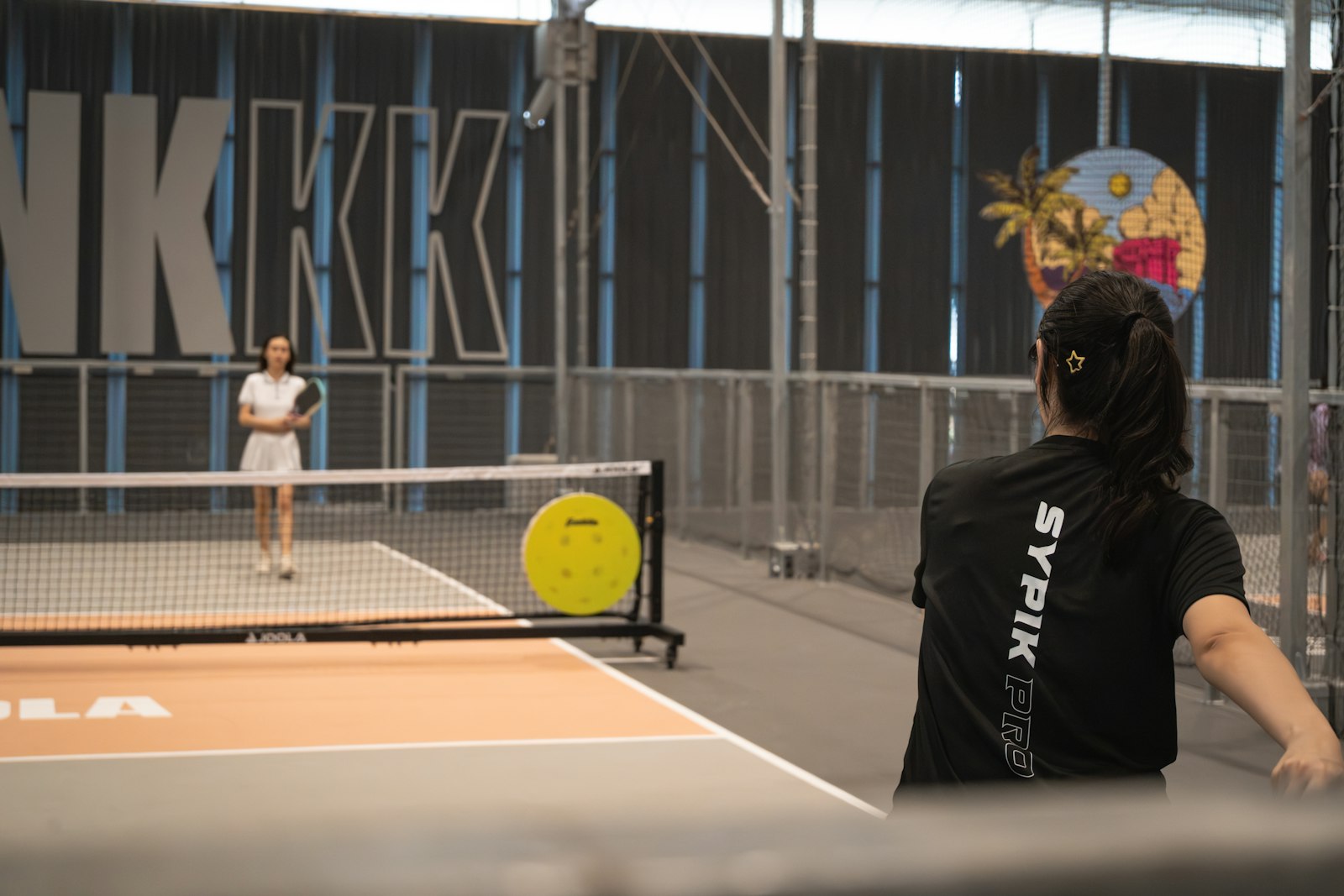Two players rally with paddles on an indoor court — pickleball, the sport this guide untangles from paddle tennis, platform tennis, and padel