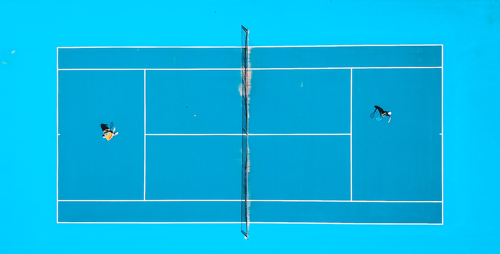 Aerial view of two players on a racket-sport court, the shared geometry that connects pickleball, tennis, and padel
