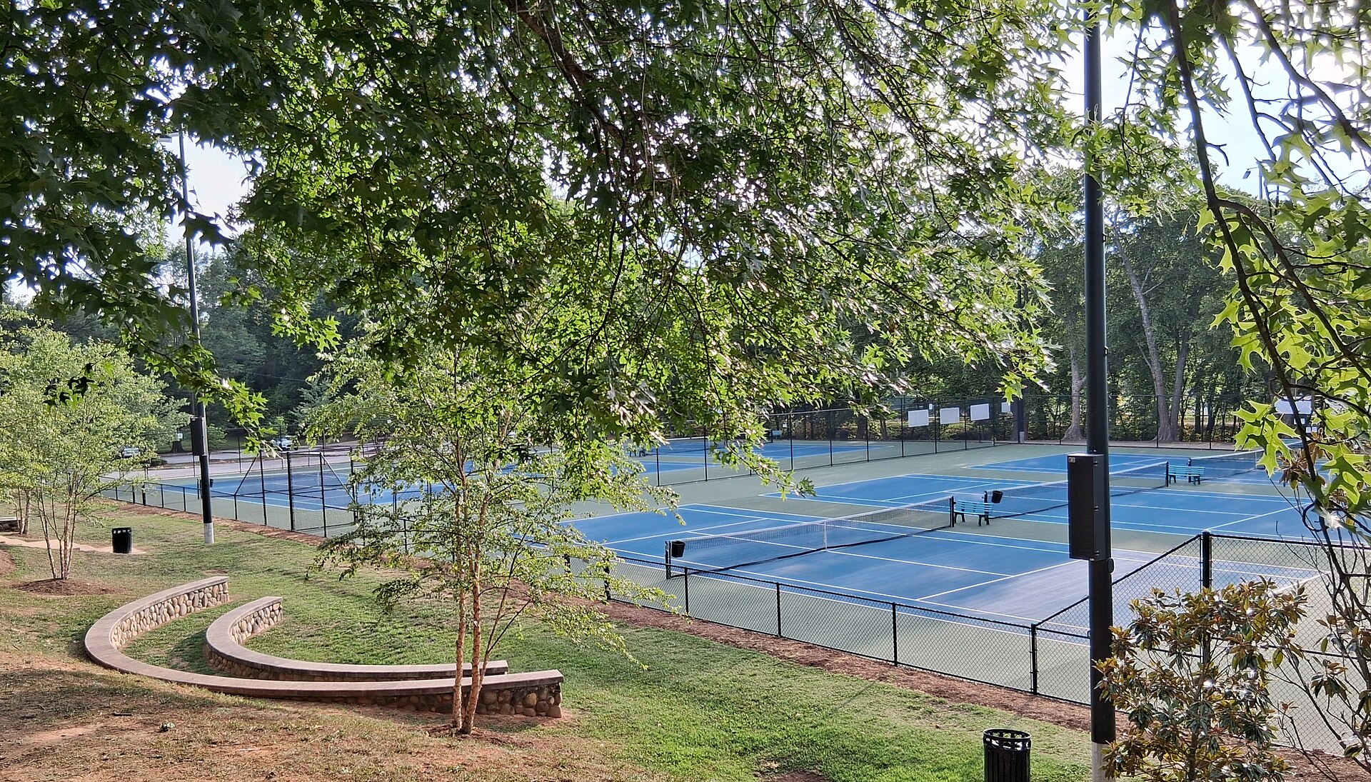 Several blue hard tennis courts with nets, lights and benches inside a fenced public park in Winston-Salem, North Carolina, seen through tree branches