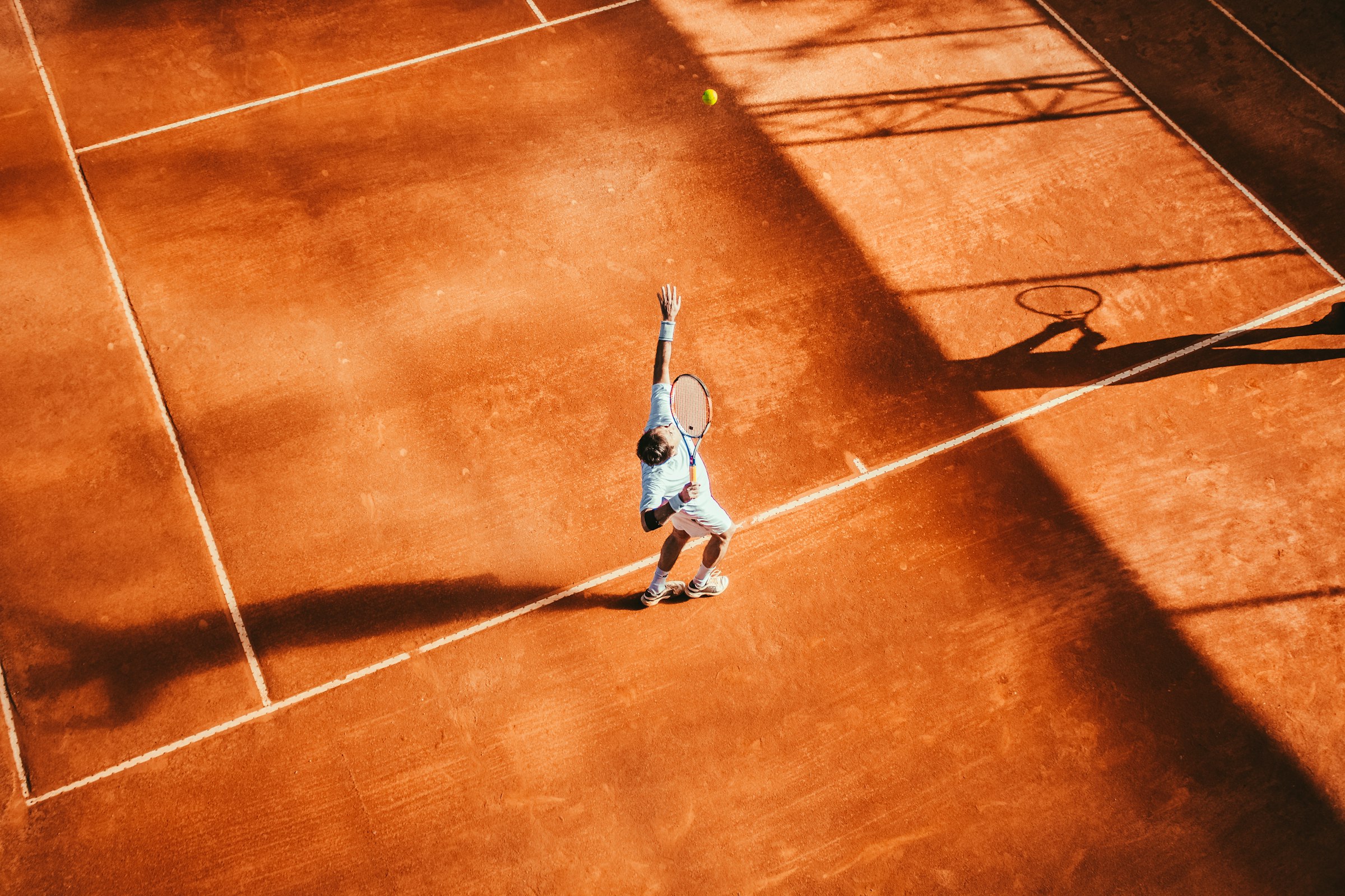 Aerial view of a lone tennis player serving on an outdoor clay court, no umpire chair or officials visible — most recreational tennis is entirely self-officiated