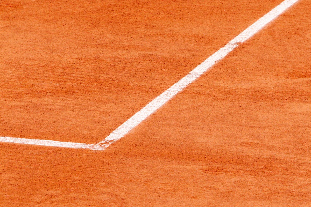 Primer plano de la línea de fondo y la línea lateral de una pista de tenis encontrándose en una esquina sobre tierra batida roja, el tipo de línea contra la que se juzga el bote de una pelota