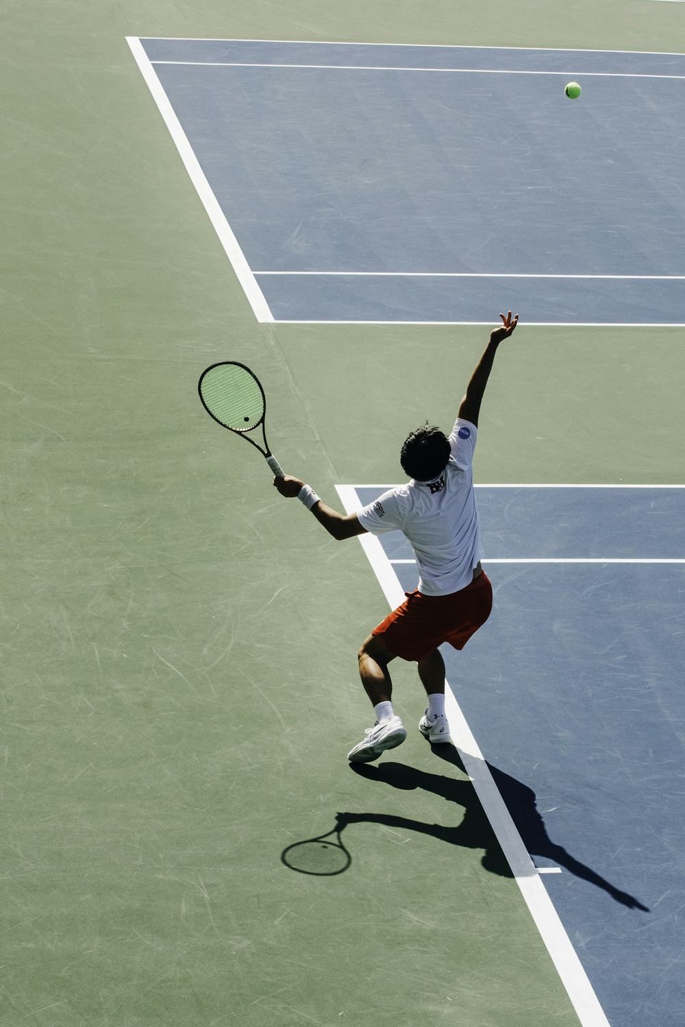 Aerial view of a tennis player at the top of their serve motion, racket extended overhead toward a ball in flight above the service box
