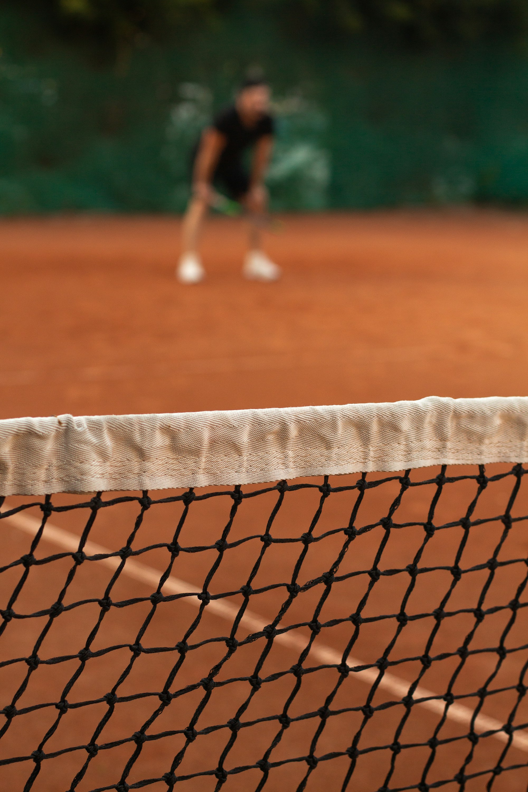 A tennis net's white tape and black mesh in sharp focus in the foreground, with a player blurred behind it crouched low in a ready position on a clay court