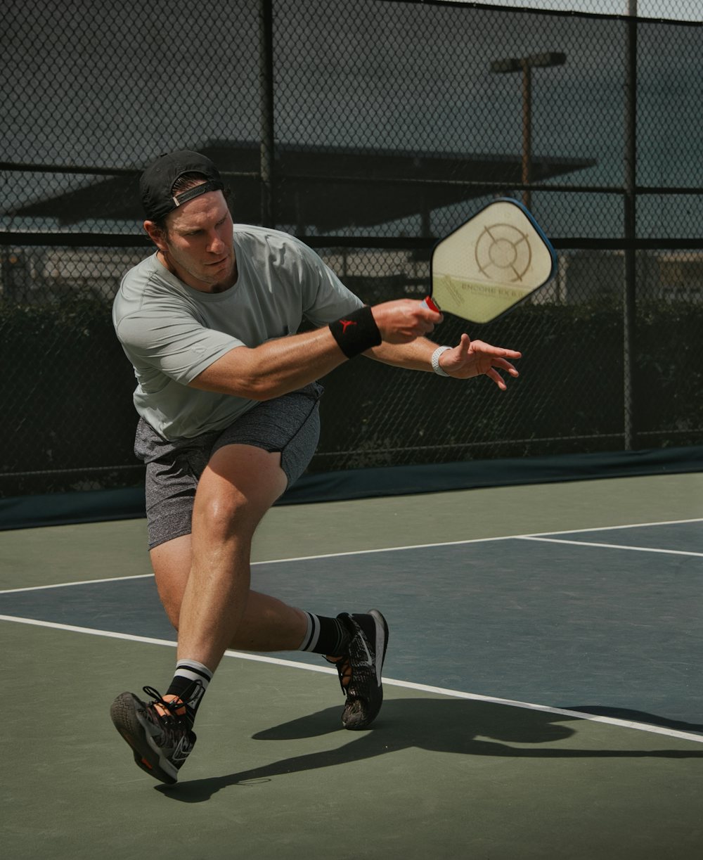 A pickleball player mid-forehand on an outdoor court, wearing a moisture-wicking t-shirt, athletic shorts, a wristband, backwards cap, and court shoes